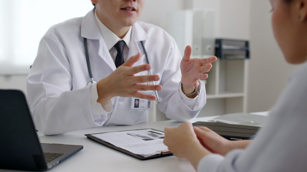Ein Arzt im Gespräch mit seinem Patienten, auf dem Tisch liegt eine Mappe mit Diagnosen, der Arzt wirkt freundlich zugewandt, die Person hört aufmerksam zu.