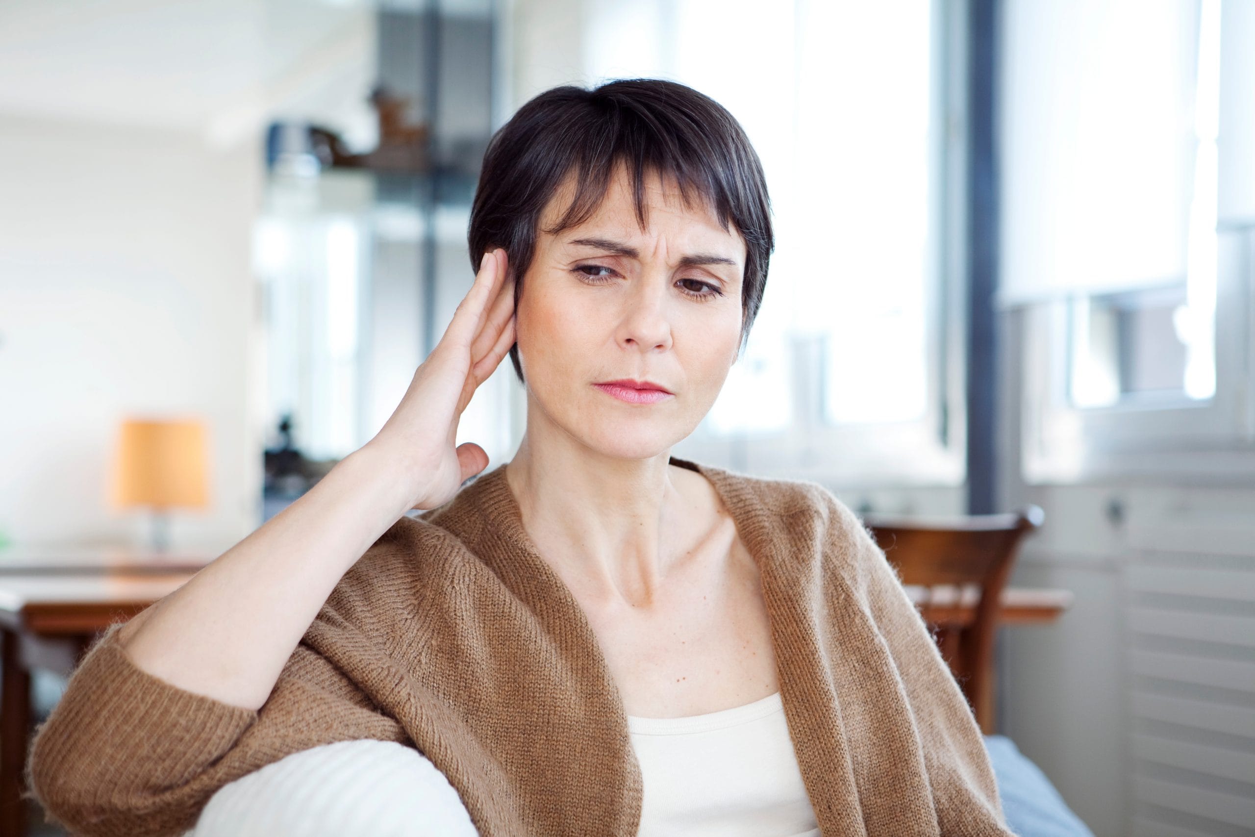 Eine Frau mittleren Alters sitzt nachdenklich auf einem Sofa und hält sich eine Hand an den Kopf, was möglicherweise auf Kopfschmerzen, Stress oder Konzentrationsprobleme hinweist.