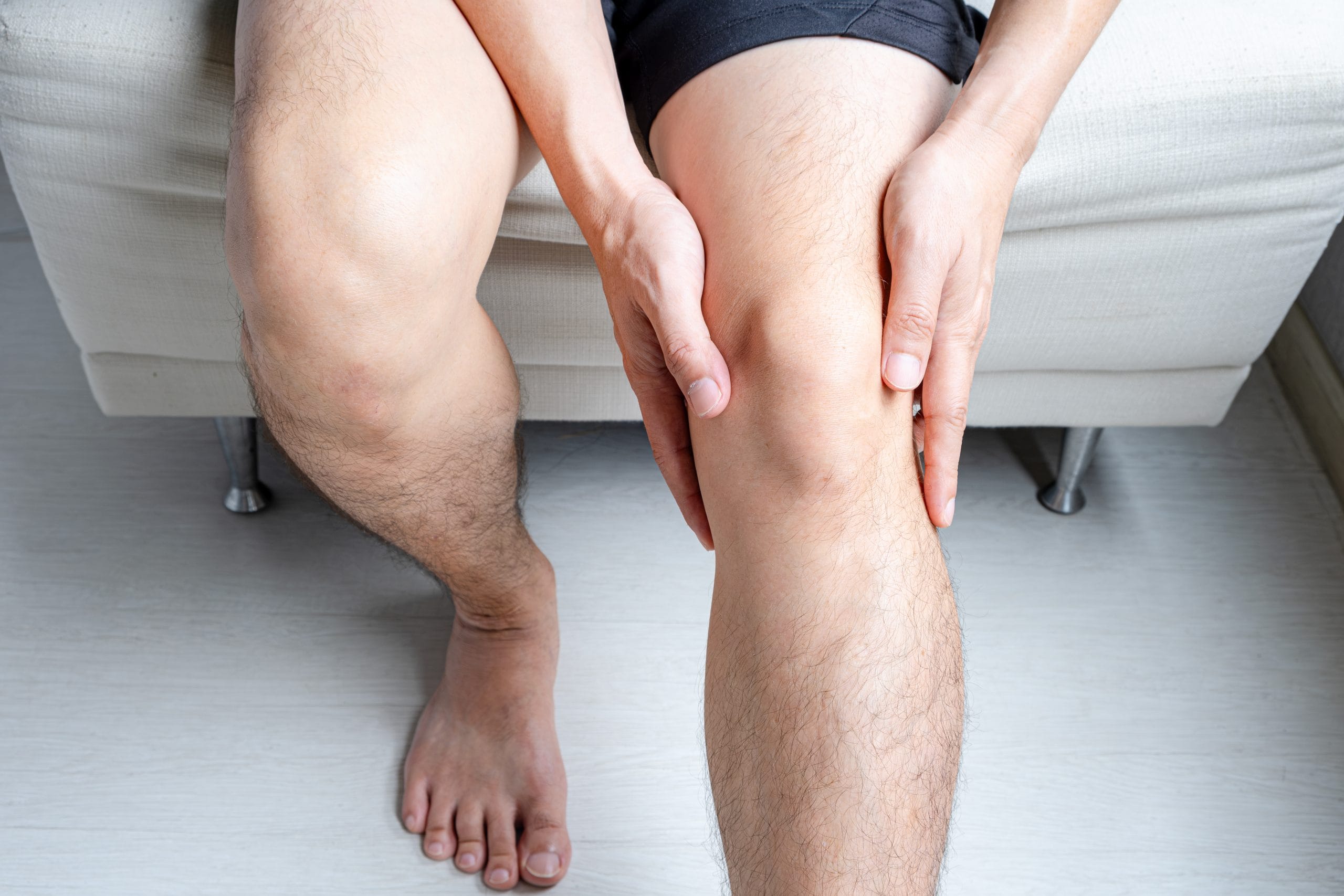 Person sitzt auf einem Sofa und hält sich das schmerzende Knie, möglicherweise aufgrund von Arthrose oder Gelenkschmerzen. Das Bild zeigt die Beine der Person und die Hände, die das Knie umfassen, und vermittelt den Eindruck von Unbehagen und Schmerz. Die Szene deutet auf die Notwendigkeit einer medizinischen Behandlung oder Schmerzlinderung hin.