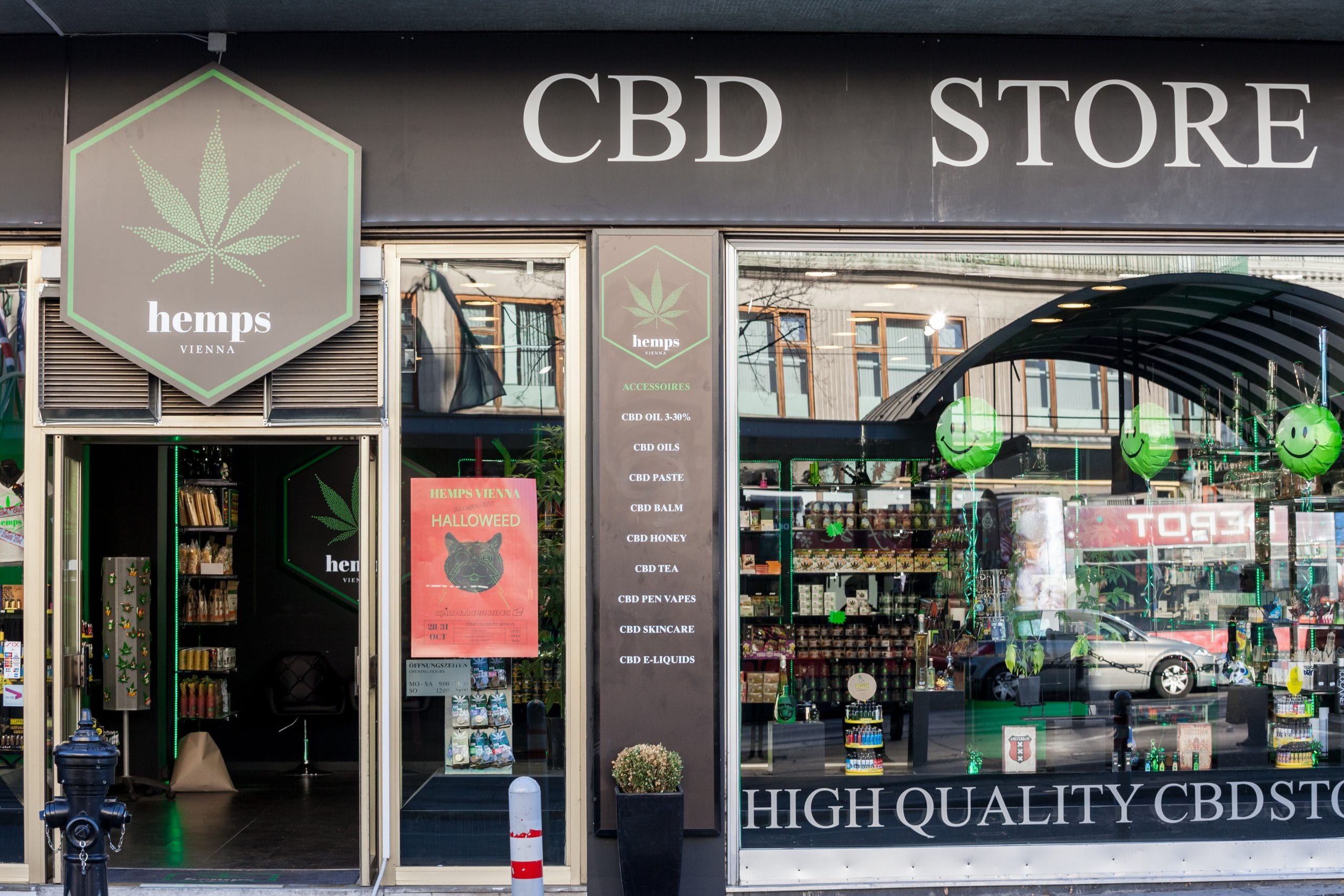 Außenansicht eines CBD-Shops mit der Aufschrift 'CBD STORE' und einer großen grünen Hanfblatt-Grafik auf einem sechseckigen Schild. Der Shop zeigt durch die Schaufenster eine Auswahl an CBD-Produkten und ist ansprechend dekoriert.