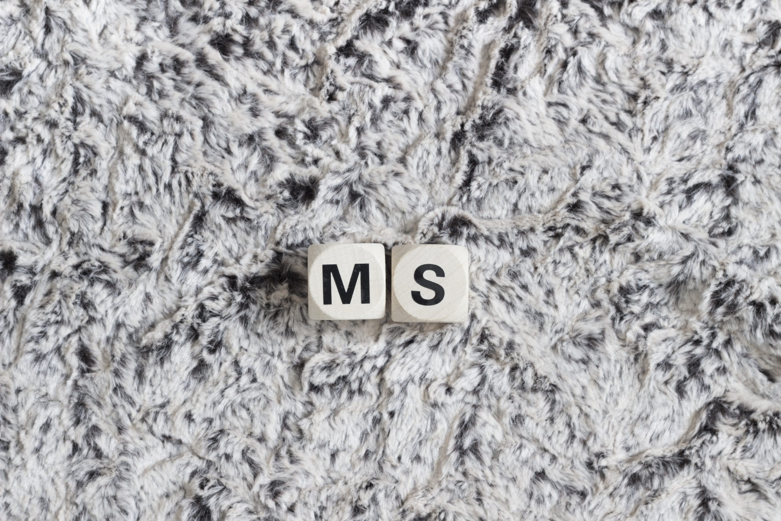 Holzwürfel mit den Buchstaben 'MS' auf einer flauschigen, grauen Oberfläche, die für Multiple Sklerose steht.