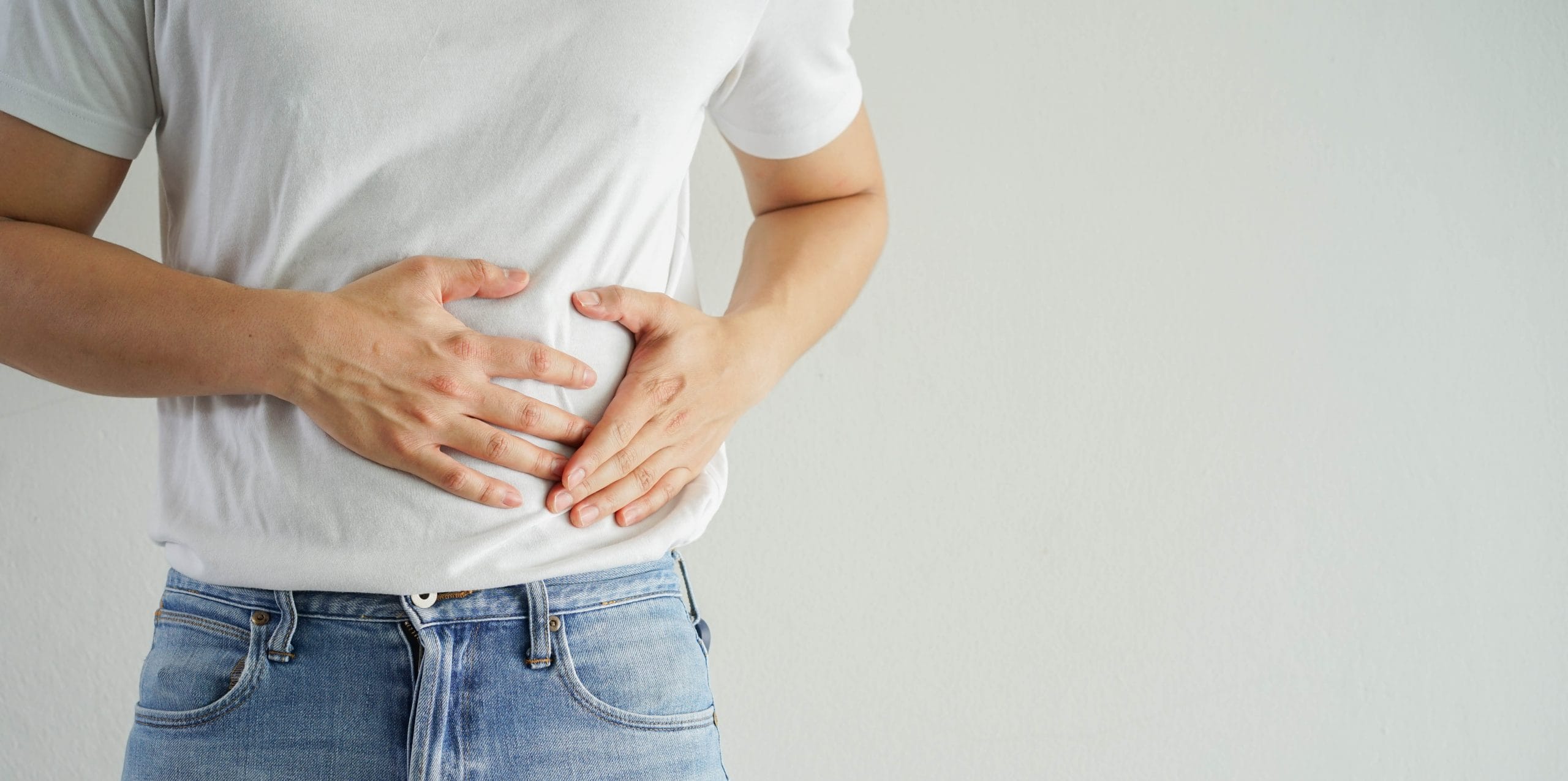 Nahaufnahme eines Mannes in Jeans und weißem T-Shirt, der sich den Bauch hält, was auf Bauchschmerzen oder Beschwerden im Verdauungstrakt hindeutet.