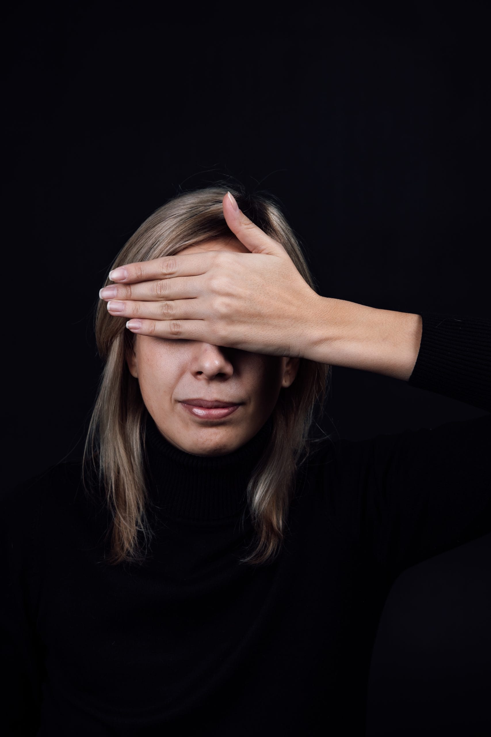 Eine Frau mit blonden Haaren, die einen schwarzen Rollkragenpullover trägt, hält sich eine Hand über ein Auge. Der Hintergrund ist dunkel.