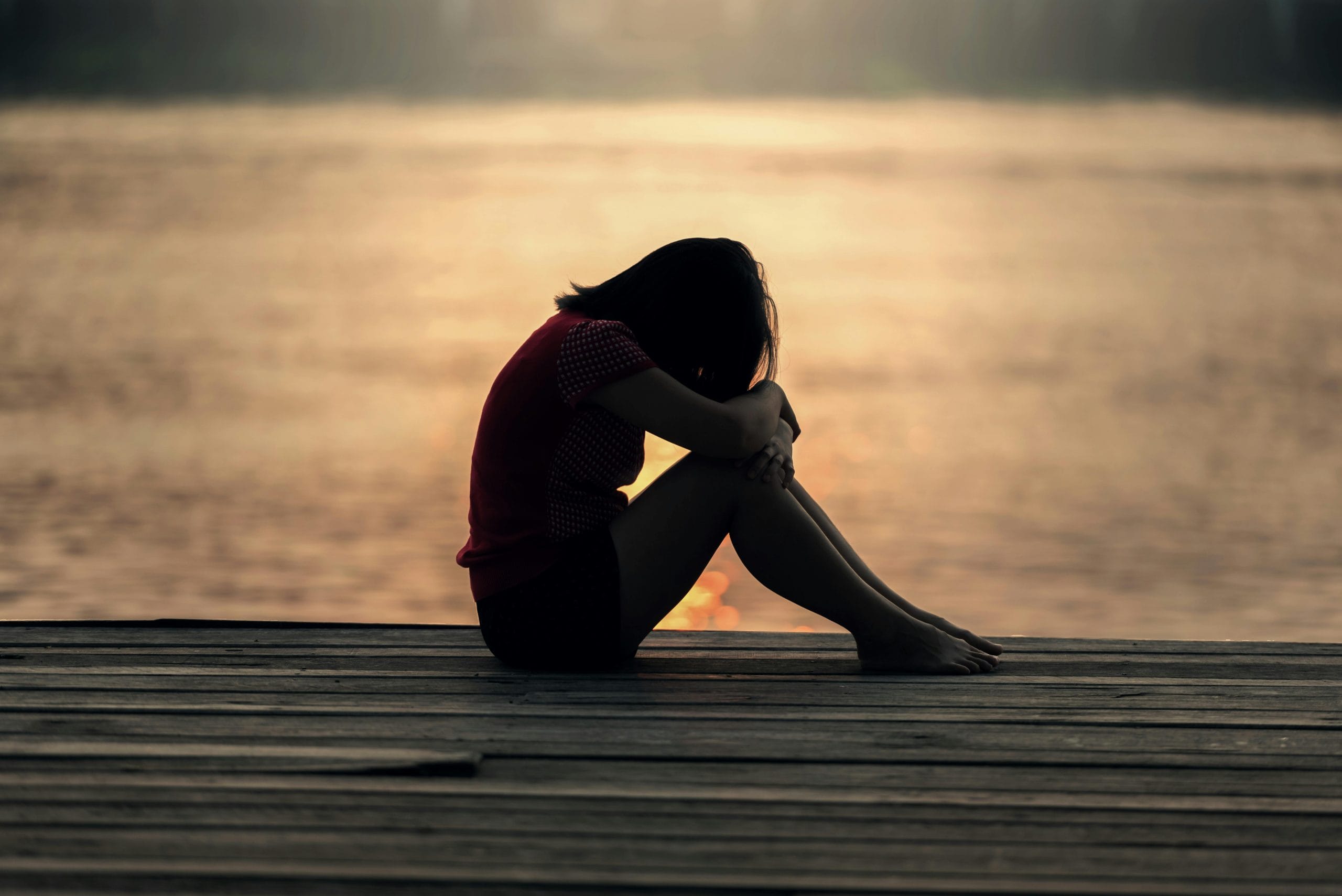 Eine Frau sitzt allein auf einem Holzsteg am Wasser, den Kopf auf die Knie gestützt. Die Abendsonne spiegelt sich im ruhigen Wasser und erzeugt eine melancholische und nachdenkliche Stimmung.