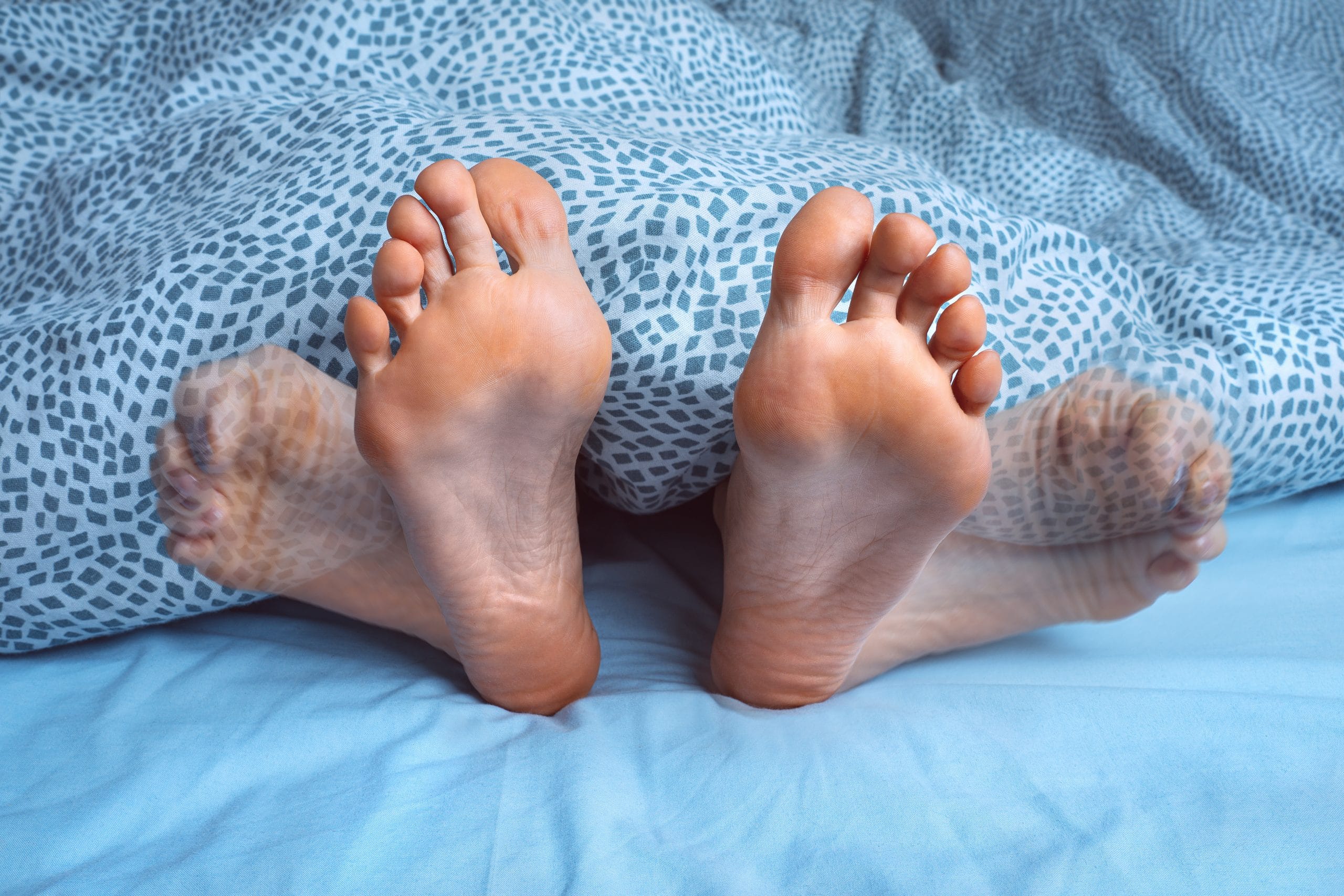 Das Bild zeigt ein Paar Füße unter einer Decke, wobei eines der Beine in Bewegung ist – ein typisches Symptom des Restless-Legs-Syndroms (RLS). Menschen mit RLS verspüren oft ein unangenehmes Kribbeln oder Zucken in den Beinen, das sie daran hindert, ruhig zu schlafen. Hausmittel wie Magnesium, Wechselduschen oder Lavendelöl können helfen, die Beschwerden zu lindern und die Schlafqualität zu verbessern.