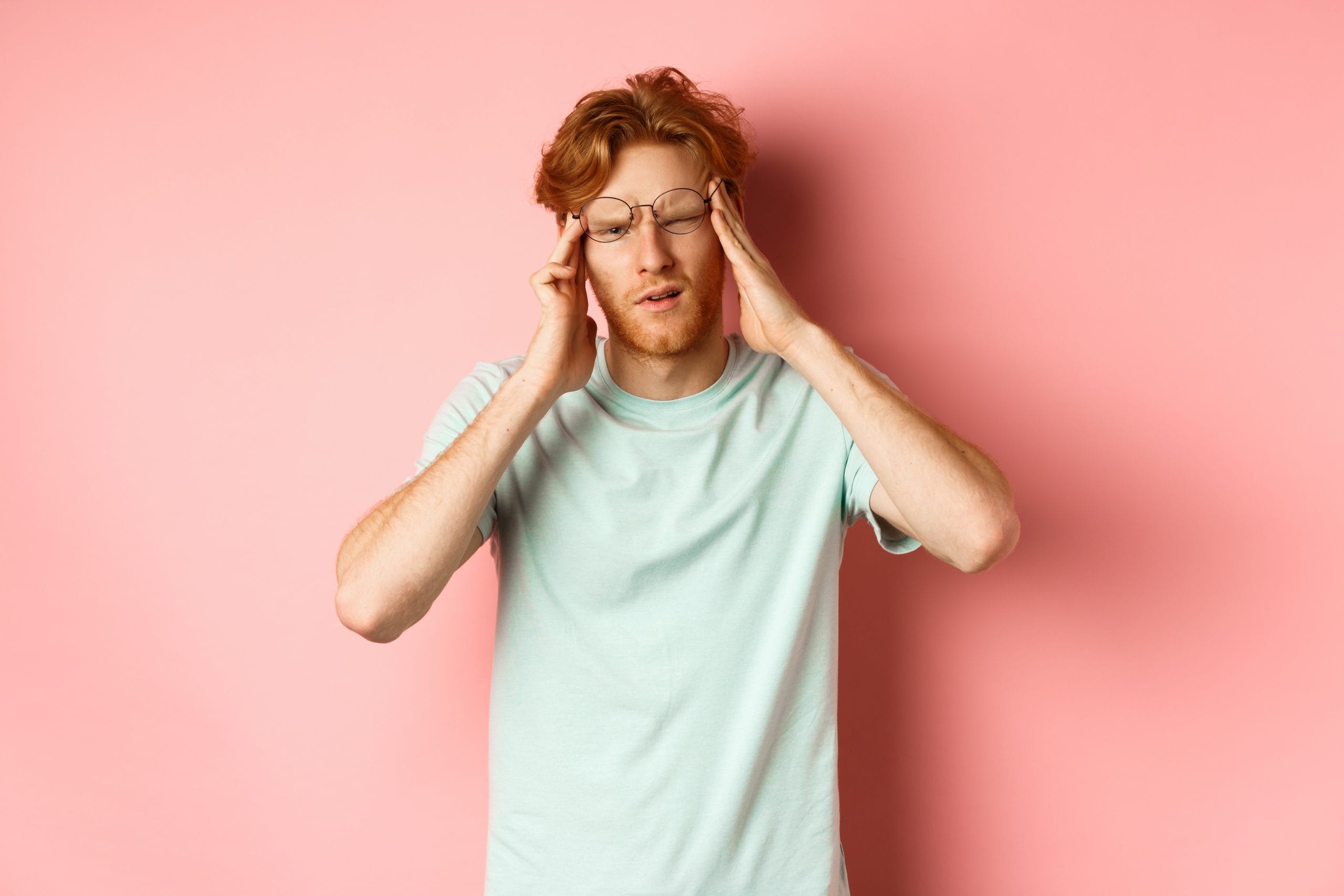 Ein junger Mann mit roten Haaren und Brille hält sich den Kopf mit beiden Händen und kneift ein Auge zusammen. Er sieht benommen aus, als hätte er mit Schwindel oder Kopfschmerzen zu kämpfen. Der Hintergrund ist rosa und der Mann trägt ein mintgrünes T-Shirt.